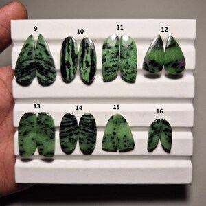Könnte beinhalten: Eine Sammlung polierter Zoisit-Edelsteine in verschiedenen Formen, darunter Tropfen und Oval. Die Steine weisen eine lebendige grüne Farbe mit schwarzen Adern auf, präsentiert auf einem weißen Tablett. Jedes Paar ist nummeriert.