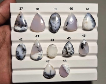 May include: A selection of polished gemstones, each with unique patterns and colours. The stones are displayed on a white, tiered surface. The gemstones vary in shape, including oval, teardrop, and triangular, with a range of colours from white to grey and lavender.