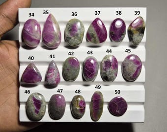 Puede incluir: Una colección de piedras preciosas de rubí pulidas en varias formas, incluyendo ovaladas, en forma de lágrima y redondas. Las piedras preciosas muestran una gama de colores que van del rojo intenso al rosa, con algunas que muestran inclusiones grises y verdes. Las piedras están dispuestas en una exhibición blanca.
