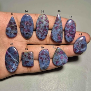 May include: A selection of polished gemstones, displaying a mottled pattern of blue and purple hues. The stones are presented in diverse shapes, such as oval, teardrop, and rectangular. Each gemstone is labelled with a number.