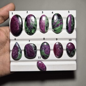 May include: A collection of eleven ruby zoisite gemstones in various shapes, including oval, teardrop, and rectangular. The stones display a mix of deep magenta and green hues, with a polished, glossy finish. The gemstones are arranged on a white display.