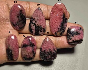 May include: A collection of polished rhodochrosite gemstones in various shapes, including oval, teardrop, and rectangular. The stones display a mix of pink and black hues, with a glossy finish. The gemstones are numbered for identification.