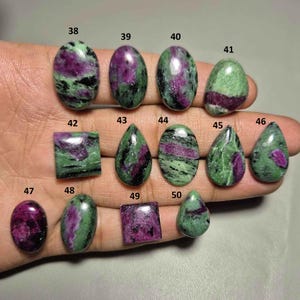 May include: A collection of polished ruby in zoisite gemstones in various shapes, including oval, teardrop, and square. The stones display a mix of deep purple and green hues, with some featuring a marbled appearance. The gemstones are numbered for identification.