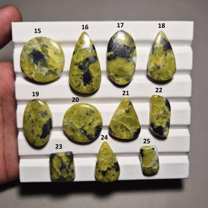 May include: A collection of polished gemstones in various shapes, including oval, teardrop, and rectangular. The stones feature a mix of yellow-green and black patterns, displayed on a white tray. The gemstones are numbered from 15 to 25.