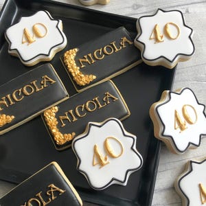 Peut inclure: Une collection de biscuits décorés sur un plateau noir. Certains biscuits sont des carrés blancs à bords festonnés avec le nombre 40 en doré. D'autres sont des rectangles noirs avec le nom NICOLA en lettres dorées et des paillettes dorées.