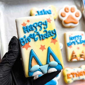 以下が含まれることがあります： 黄色いアイシング、オレンジ色の星、そして青い文字で「Happy Birthday」と書かれ、青とオレンジのキャラクターの顔が描かれた長方形のクッキー。金属製のトレイには、足跡や骨の形をした他のクッキーも並んでいる。