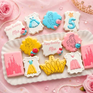 Puede incluir: Una colección de galletas decoradas en una bandeja blanca, con diseños de princesas que incluyen vestidos, castillos, una corona y siluetas. Algunas galletas muestran el texto Happy Birthday Mia y el número 5 con una varita.