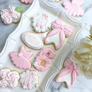May include: A collection of decorated sugar cookies in various shapes, including ballet shoes, bows, and a number 6. The cookies are primarily pink and white, with floral and ballet-themed designs. One cookie is inscribed with the name "Elise".