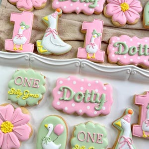 Può includere: Una collezione di biscotti di zucchero decorati, con una forma a "1" con un'oca, un biscotto a forma di oca e biscotti con il nome "Dotty" e le parole "One Silly Goose". I biscotti sono decorati con glassa rosa, bianca, gialla e verde.