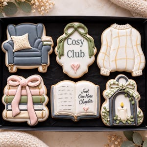 May include: A set of six decorated sugar cookies in a black box. Shapes include a blue armchair, a plaque with the text Cosy Club, a white sweater, a stack of books with a pink bow, an open book with the text Just One More Chapter, and a lantern.