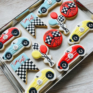 Può includere: Un set di nove biscotti decorati a tema corsa. I biscotti sono decorati con auto da corsa rosse, blu, gialle e bianche, bandiere a scacchi, palloncini e il testo "Two Fast" e "Ready Set Go!!".