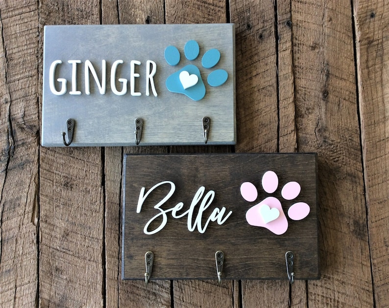 K&ouml;nnte beinhalten: Zwei Schl&uuml;sselhaken aus Holz mit Pfotenabdruck-Designs und einem Herz. Der obere Haken ist grau mit dem Namen "Ginger" und einem t&uuml;rkisfarbenen Pfotenabdruck. Der untere Haken ist braun mit dem Namen "Bella" und einem rosafarbenen Pfotenabdruck.