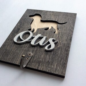 May include: A wooden sign with a cutout of a dachshund dog and the name "Otis" in white lettering. The sign has a metal hook attached to the bottom.