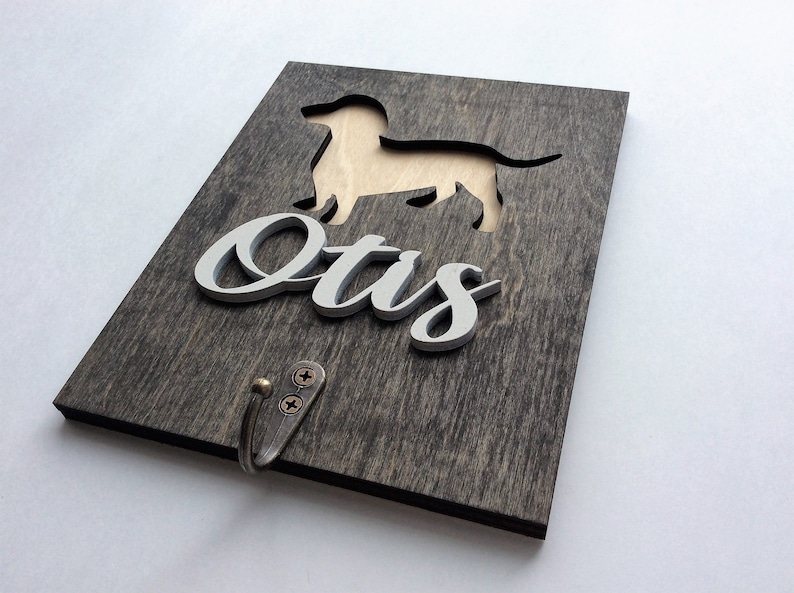 May include: A wooden sign with a cutout of a dachshund dog and the name "Otis" in white lettering. The sign has a metal hook attached to the bottom.