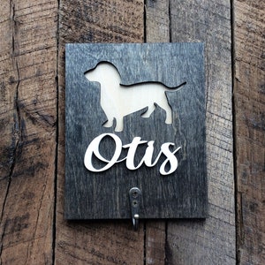 May include: A dark wood sign with a white dachshund silhouette and the name "Otis" in white lettering. The sign has a metal hook on the bottom.