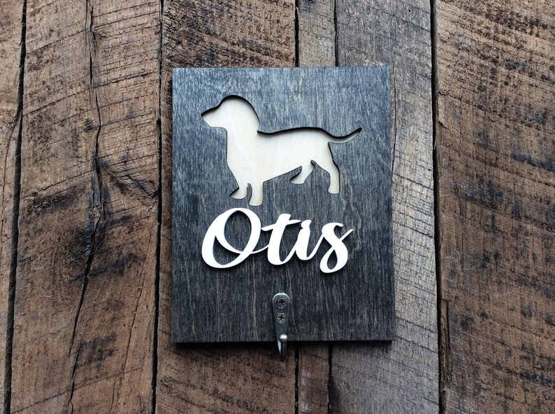 May include: A dark wood sign with a white dachshund silhouette and the name "Otis" in white lettering. The sign has a metal hook on the bottom.
