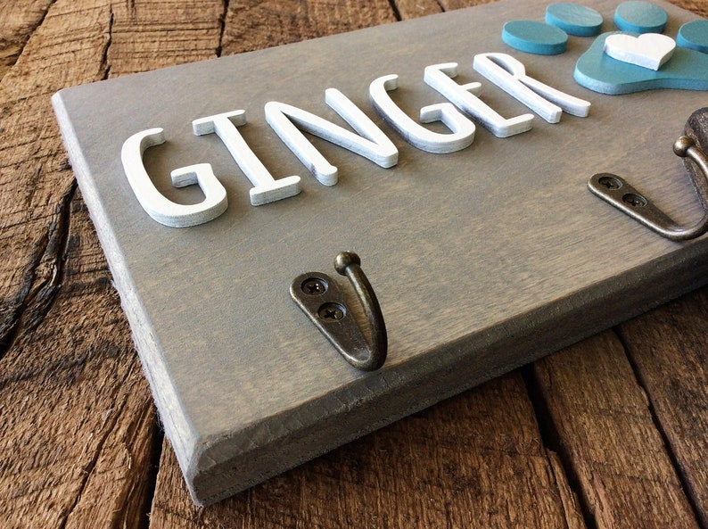 K&ouml;nnte beinhalten: Ein graues Holzschild mit dem Namen "Ginger" in wei&szlig;en Buchstaben. Das Schild hat zwei Haken und einen blauen Pfotenabdruck mit einem wei&szlig;en Herzen.