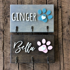 K&ouml;nnte beinhalten: Zwei Schl&uuml;sselhaken aus Holz mit Pfotenabdruck-Designs und einem Herz. Der obere Haken ist grau mit einem blauen Pfotenabdruck und dem Text "GINGER". Der untere Haken ist braun mit einem rosa Pfotenabdruck und dem Text "Bella".