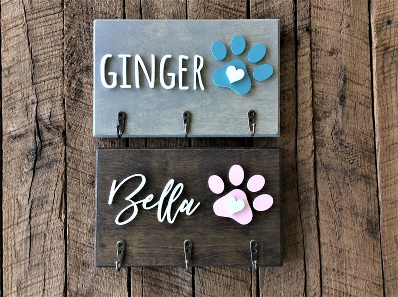 K&ouml;nnte beinhalten: Zwei Schl&uuml;sselhaken aus Holz mit Pfotenabdruck-Designs und einem Herz. Der obere Haken ist grau mit einem blauen Pfotenabdruck und dem Text "GINGER". Der untere Haken ist braun mit einem rosa Pfotenabdruck und dem Text "Bella".