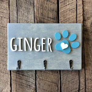K&ouml;nnte beinhalten: Ein grauer Holz-Schl&uuml;sselhalter mit dem Namen "GINGER" in wei&szlig;en Buchstaben und einem blauen Pfotenabdruck mit einem wei&szlig;en Herzen in der Mitte. Es gibt drei Haken an der Unterseite.