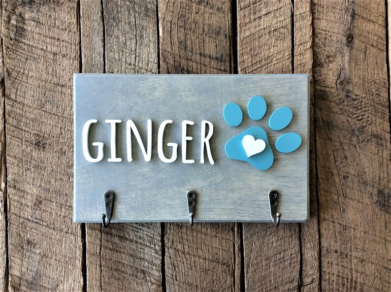 K&ouml;nnte beinhalten: Ein grauer Holz-Schl&uuml;sselhalter mit dem Namen "GINGER" in wei&szlig;en Buchstaben und einem blauen Pfotenabdruck mit einem wei&szlig;en Herzen in der Mitte. Es gibt drei Haken an der Unterseite.