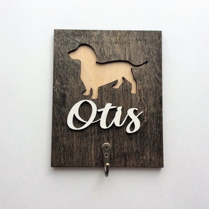 May include: A wooden sign with a cutout of a dachshund and the name "Otis" written in white. The sign has a hook for hanging.