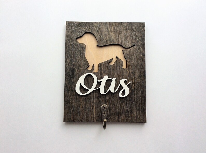 May include: A wooden sign with a cutout of a dachshund and the name "Otis" written in white. The sign has a hook for hanging.
