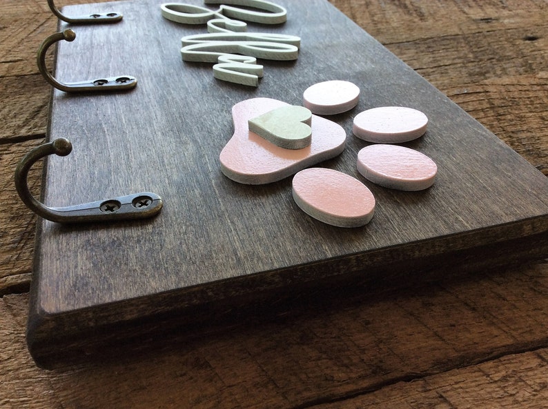 K&ouml;nnte beinhalten: Holz-Schl&uuml;sselhalter mit drei Haken, dem Wort "Willkommen" in wei&szlig;en Buchstaben und einem rosa Pfotenabdruck mit einem kleinen Herz in der Mitte.