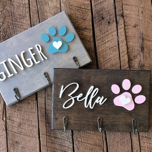 K&ouml;nnte beinhalten: Zwei h&ouml;lzerne Hunde-Leinenhalter mit Pfotenabdruck-Designs. Der obere Halter ist grau mit einem t&uuml;rkisfarbenen Pfotenabdruck und dem Text "GINGER". Der untere Halter ist braun mit einem rosafarbenen Pfotenabdruck und dem Text "Bella".