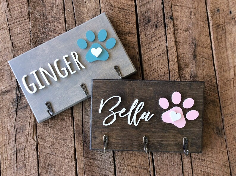 K&ouml;nnte beinhalten: Zwei h&ouml;lzerne Hunde-Leinenhalter mit Pfotenabdruck-Designs. Der obere Halter ist grau mit einem t&uuml;rkisfarbenen Pfotenabdruck und dem Text "GINGER". Der untere Halter ist braun mit einem rosafarbenen Pfotenabdruck und dem Text "Bella".