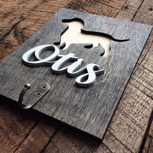May include: A wooden sign with a cutout of a dachshund dog and the name "Otis" in white lettering. The sign has a metal hook attached to the bottom.