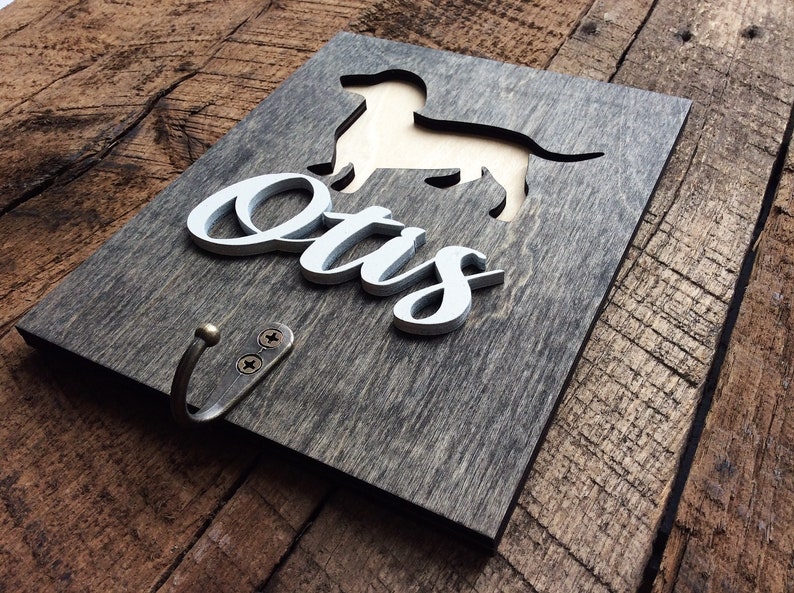 May include: A wooden sign with a cutout of a dachshund dog and the name "Otis" in white lettering. The sign has a metal hook attached to the bottom.