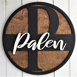 May include: A round wooden sign with a dark brown wood grain background. A black letter "P" and the name "Palen" in white script are layered on top. The sign is framed with a black border.