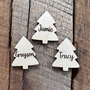 May include: Three wooden Christmas tree ornaments with names: "Jamie", "Grayson", and "Tracy" in a cursive font. The ornaments are light-colored and sit on a rustic wooden surface.