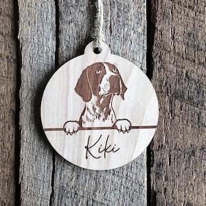 May include: A round wooden ornament with a laser-engraved image of a brown and white dog peeking over the top. The dog's name, "Kiki", is engraved below the image.
