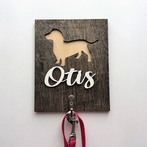 May include: A wooden dog leash hook with a dachshund silhouette cutout and the name "Otis" in white lettering. A pink leash is hanging from the hook.