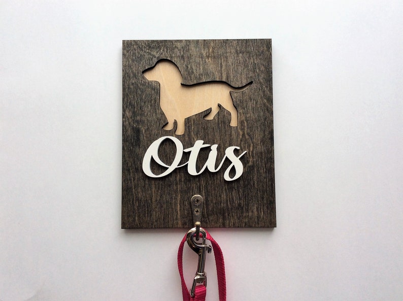May include: A wooden dog leash hook with a dachshund silhouette cutout and the name "Otis" in white lettering. A pink leash is hanging from the hook.