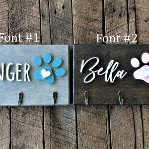 K&ouml;nnte beinhalten: Zwei h&ouml;lzerne Hunde-Leinenhalter mit Pfotenabdruck-Designs. Der Halter links ist grau mit einem t&uuml;rkisfarbenen Pfotenabdruck und dem Text "GINGER". Der Halter rechts ist braun mit einem rosa Pfotenabdruck und dem Text "Bella".