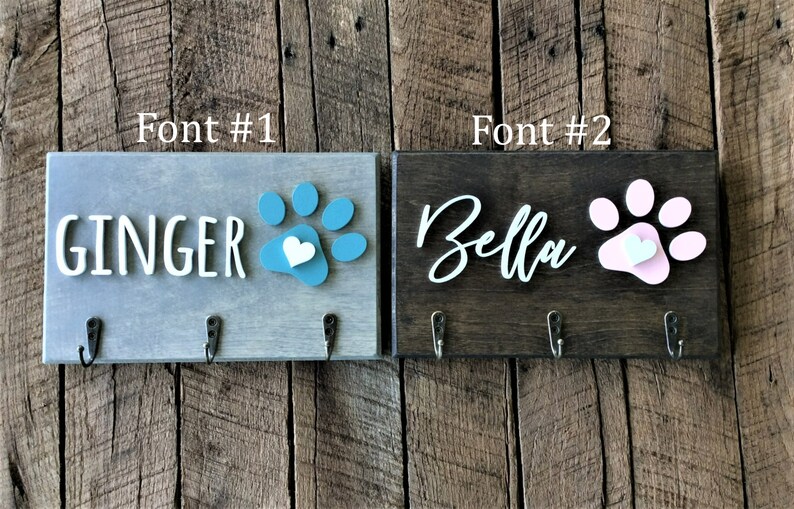 K&ouml;nnte beinhalten: Zwei h&ouml;lzerne Hunde-Leinenhalter mit Pfotenabdruck-Designs. Der Halter links ist grau mit einem t&uuml;rkisfarbenen Pfotenabdruck und dem Text "GINGER". Der Halter rechts ist braun mit einem rosa Pfotenabdruck und dem Text "Bella".