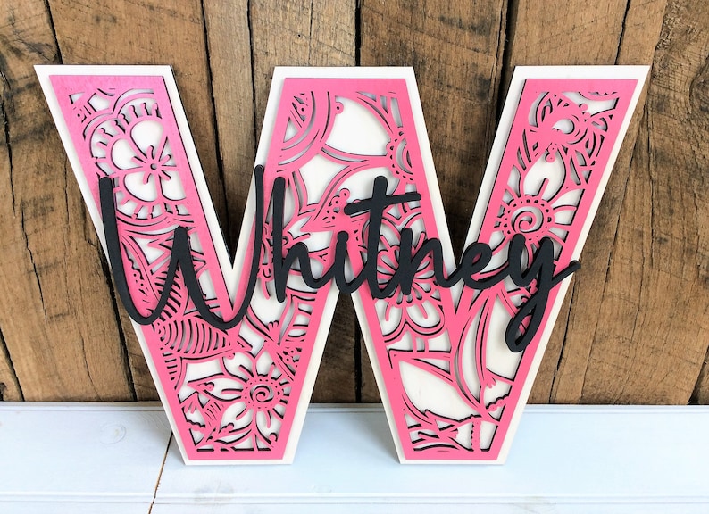 May include: Pink wooden letter W with a white background and a black floral design. The name "Whitney" is written in black cursive across the letter.