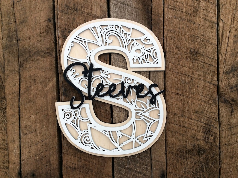 May include: A white wooden letter S with a decorative cutout design. The letter is decorated with a black cursive name "Steeves".