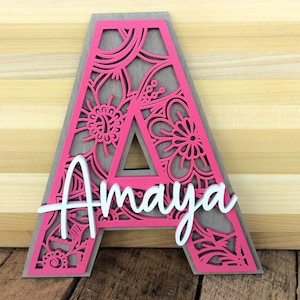May include: A pink wooden letter A with a decorative floral design. The letter is cut out and the design is visible through the cutouts. The name "Amaya" is written in white on the letter.