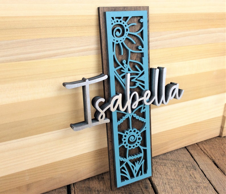 May include: A wooden sign with a blue and brown floral design and the name "Isabella" in silver lettering.