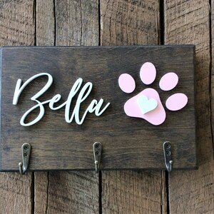K&ouml;nnte beinhalten: Ein Holzschild mit dem Namen "Bella" in wei&szlig;er Kursivschrift und einem rosa Pfotenabdruck mit einem wei&szlig;en Herzen in der Mitte. Am unteren Rand des Schildes befinden sich drei Haken.