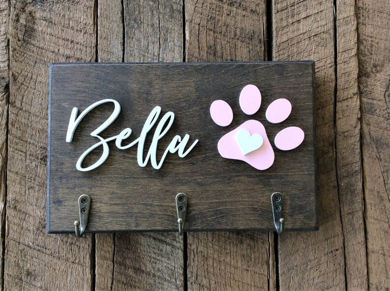 K&ouml;nnte beinhalten: Ein Holzschild mit dem Namen "Bella" in wei&szlig;er Kursivschrift und einem rosa Pfotenabdruck mit einem wei&szlig;en Herzen in der Mitte. Am unteren Rand des Schildes befinden sich drei Haken.
