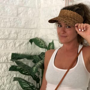 May include: A woven straw visor with a curved brim. The visor is a light brown colour and has a textured appearance. The word "TROPICS" is visible in the background. The visor is a sun protection accessory.