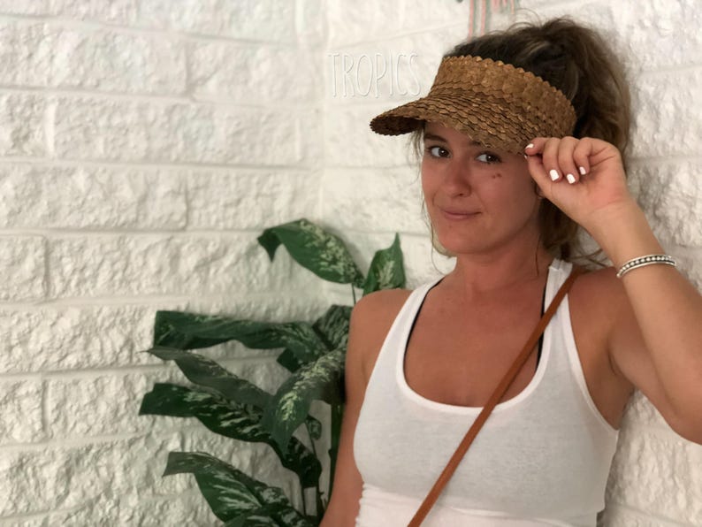 May include: A woven straw visor with a curved brim. The visor is a light brown colour and has a textured appearance. The word "TROPICS" is visible in the background. The visor is a sun protection accessory.