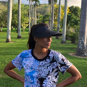 May include: A black woven visor and a short-sleeved shirt with a floral and leaf pattern. The shirt has a blue and black colour scheme with the word "JOLYN" printed on the front. The background is a green field with tall trees.