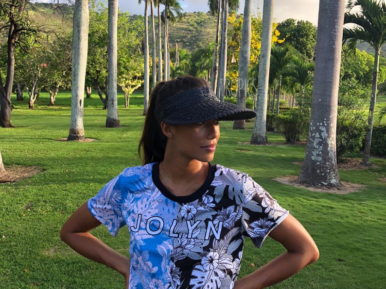 May include: A black woven visor and a short-sleeved shirt with a floral and leaf pattern. The shirt has a blue and black colour scheme with the word "JOLYN" printed on the front. The background is a green field with tall trees.