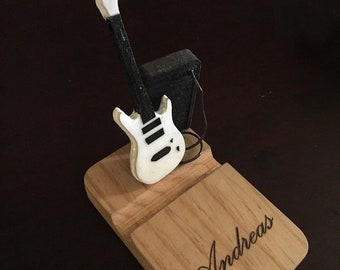 May include: A wooden phone stand shaped like a guitar, with the name "Andreas" engraved on the base. It features a miniature white and black guitar and a small black amplifier. The stand is made of light-coloured wood.